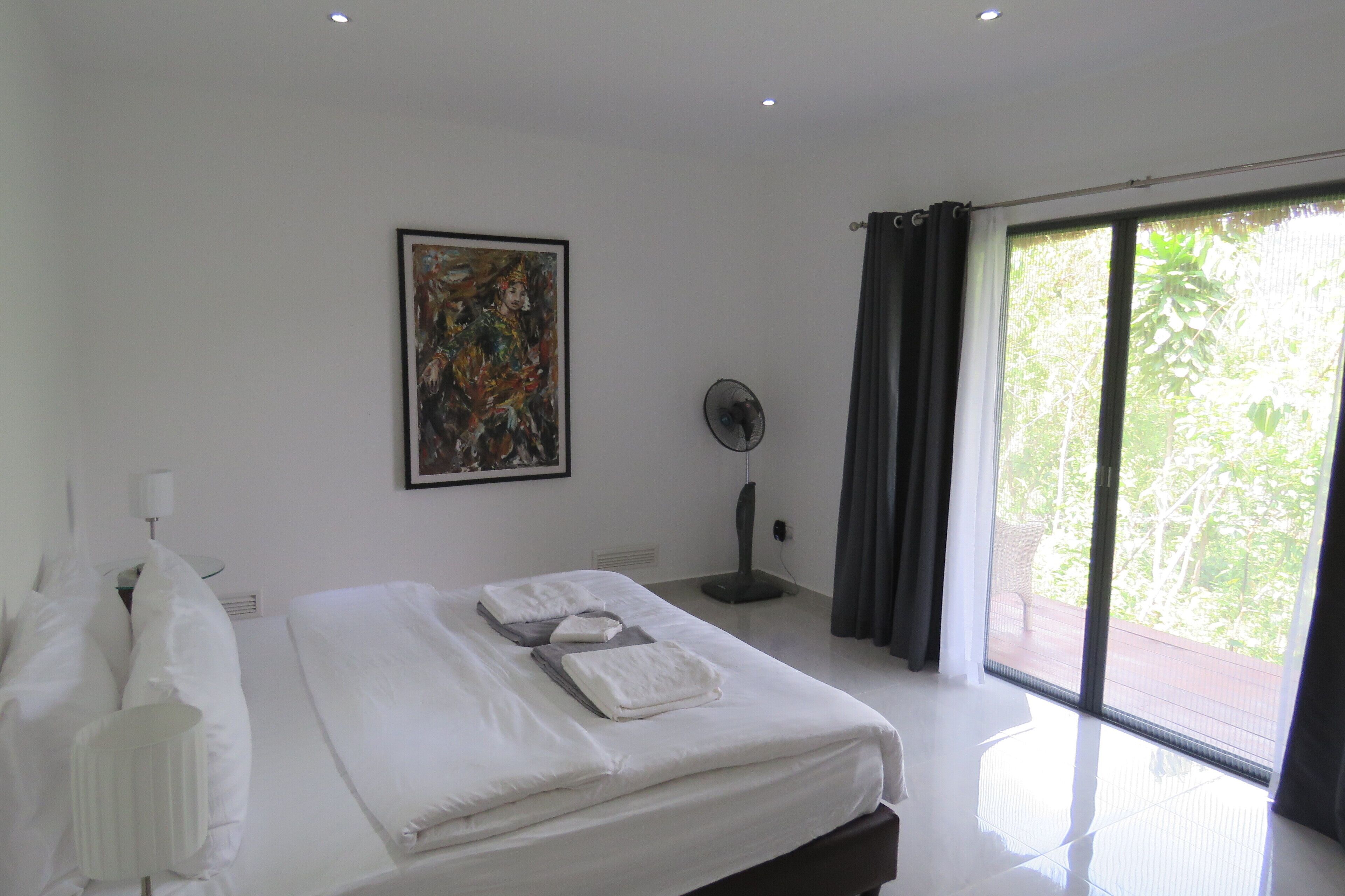 A modern hotel room with a large bed, a ceiling fan, a window with curtains, and a painting on the wall.