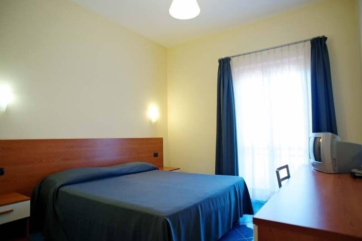A hotel room with a single bed, a wooden headboard, a desk with a television, and a window with curtains.