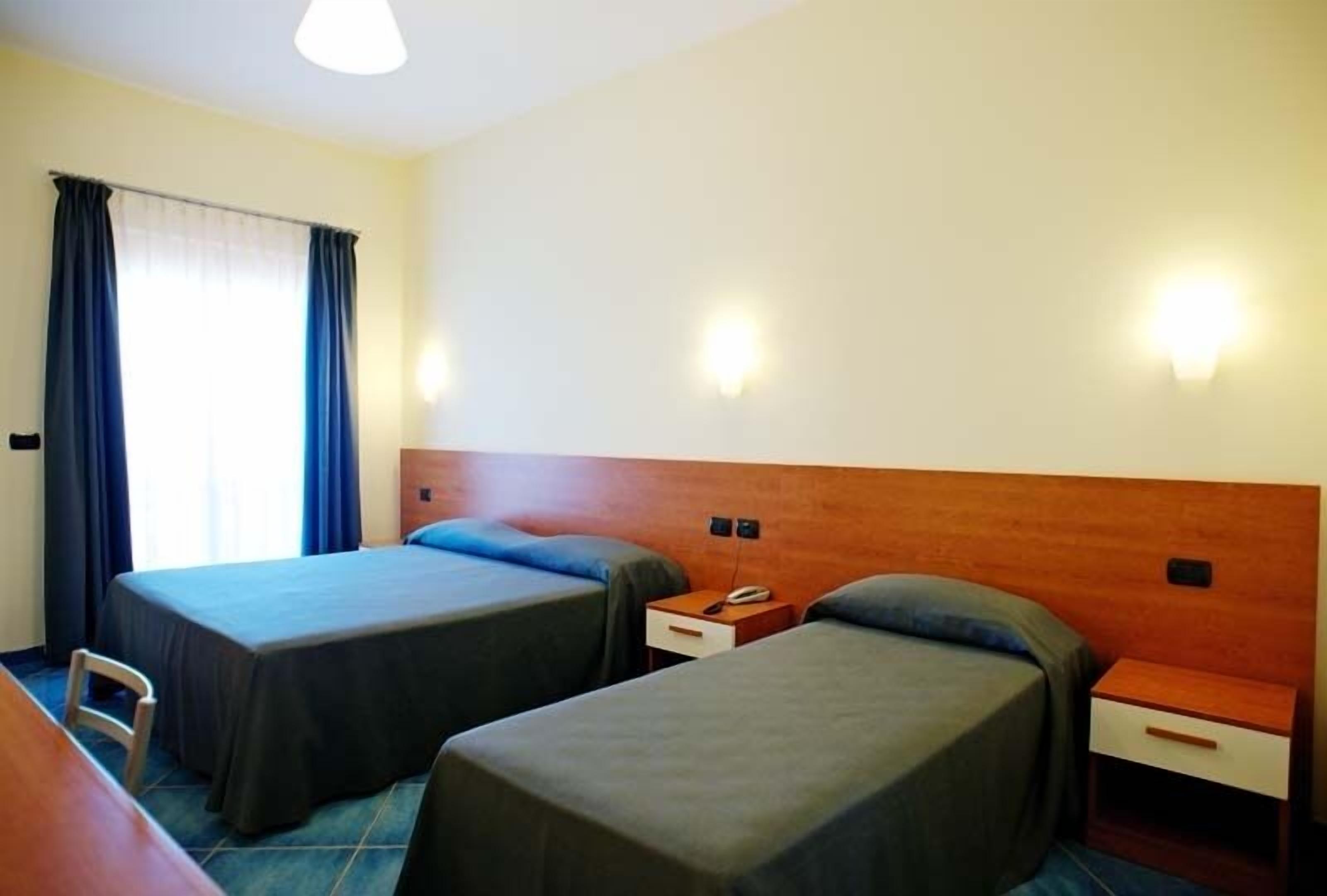 A hotel room with two single beds, a wooden headboard, bedside tables with lamps, and a window with curtains.