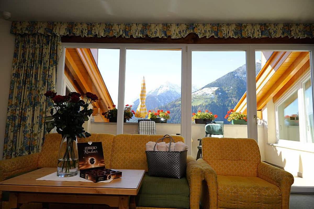 Un salon chaleureux avec un canapé, une table ornée d’un vase de fleurs et une vue sur les montagnes, visible à travers de grandes fenêtres.