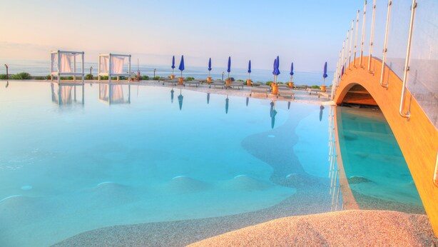 A clear swimming pool with a bridge and umbrellas, surrounded by a pebble beach and a clear sky.