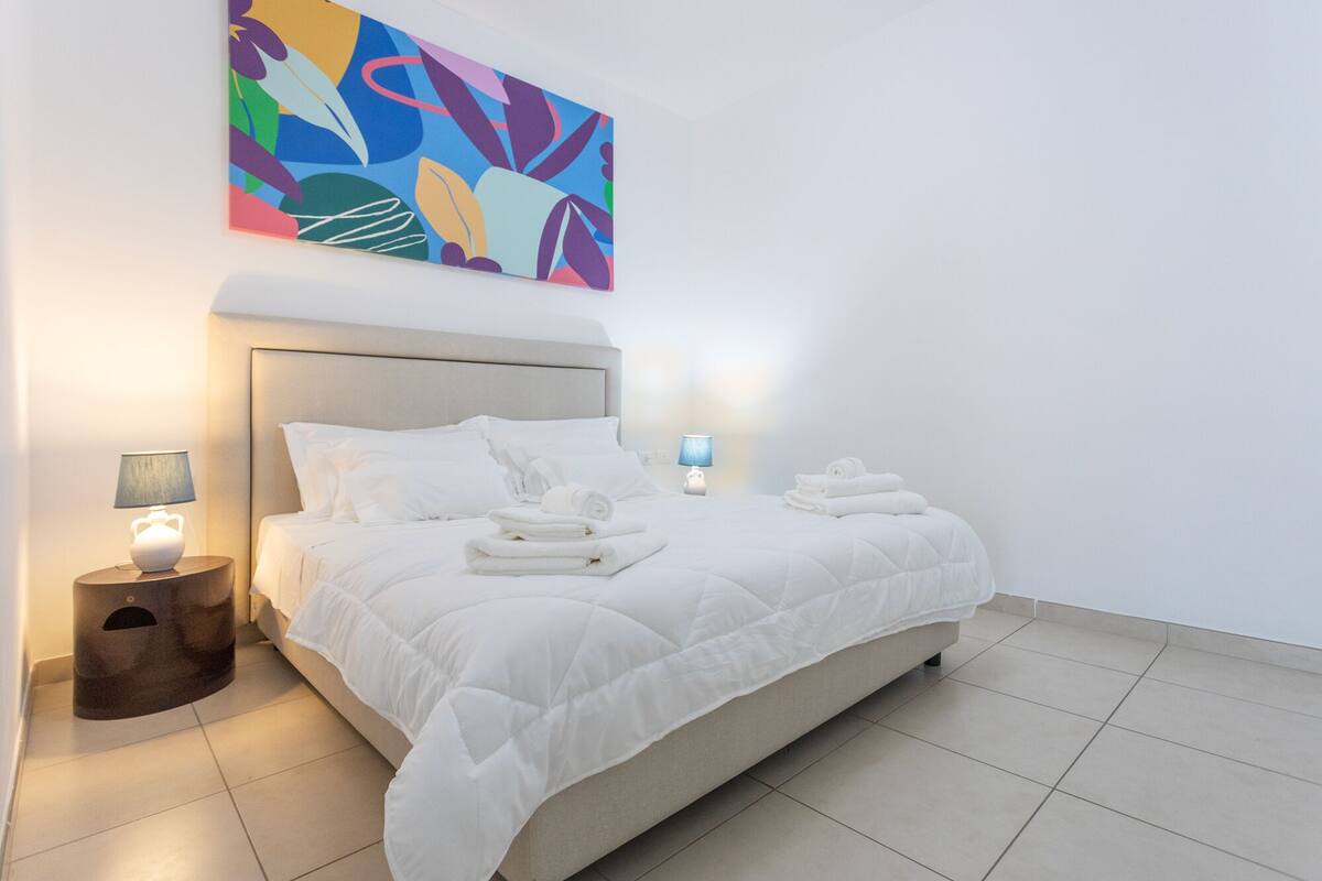 A neatly made bed with white bedding, two bedside lamps, and a colorful abstract painting on the wall.