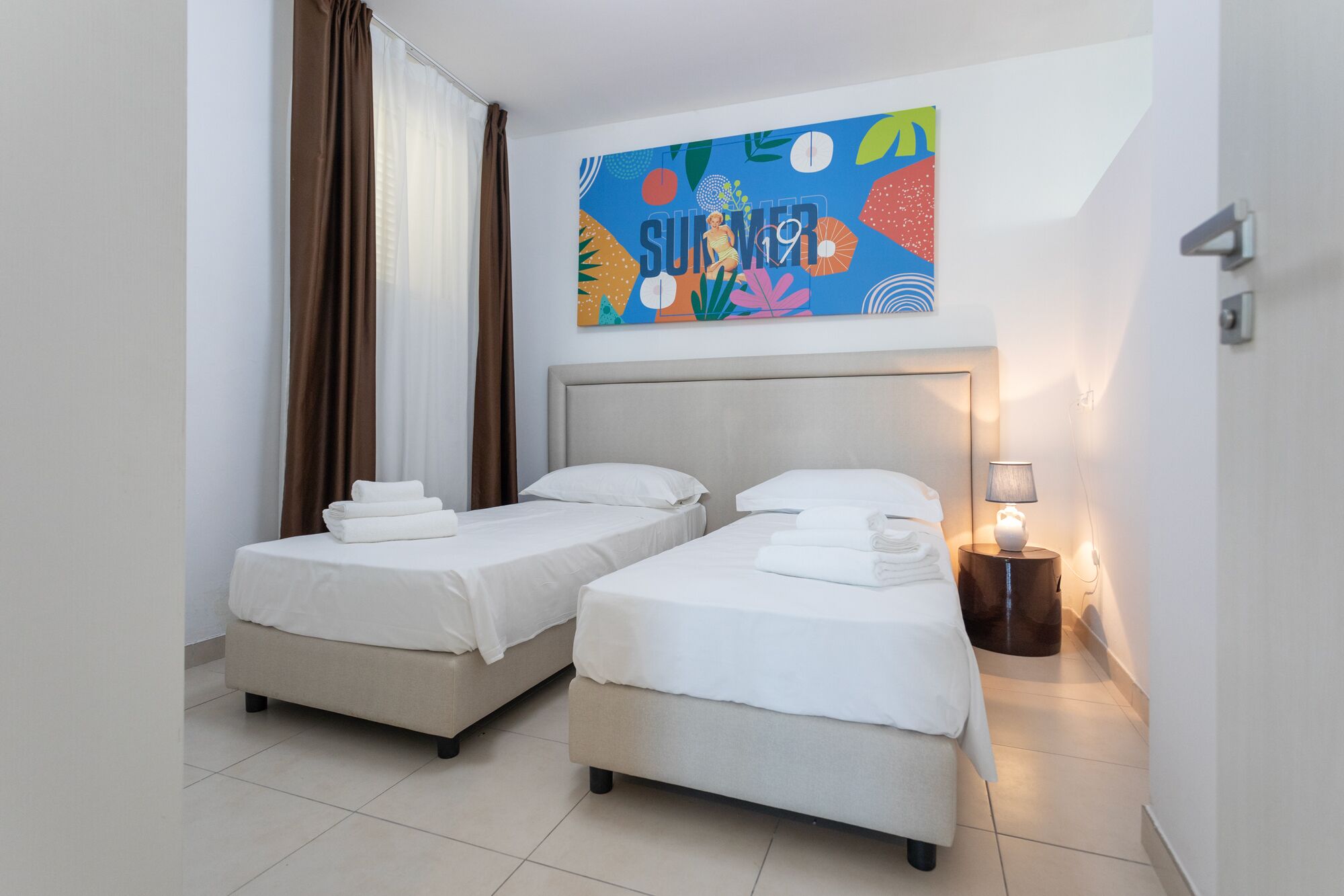 A hotel room with two beds, a nightstand with a lamp, and a colorful wall decoration.