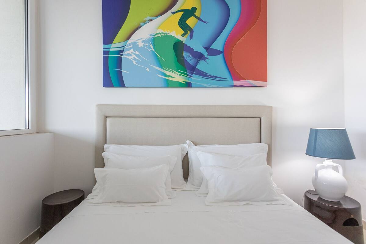 A neatly made bed with white linens and pillows, a bedside lamp, and a colorful abstract painting on the wall.