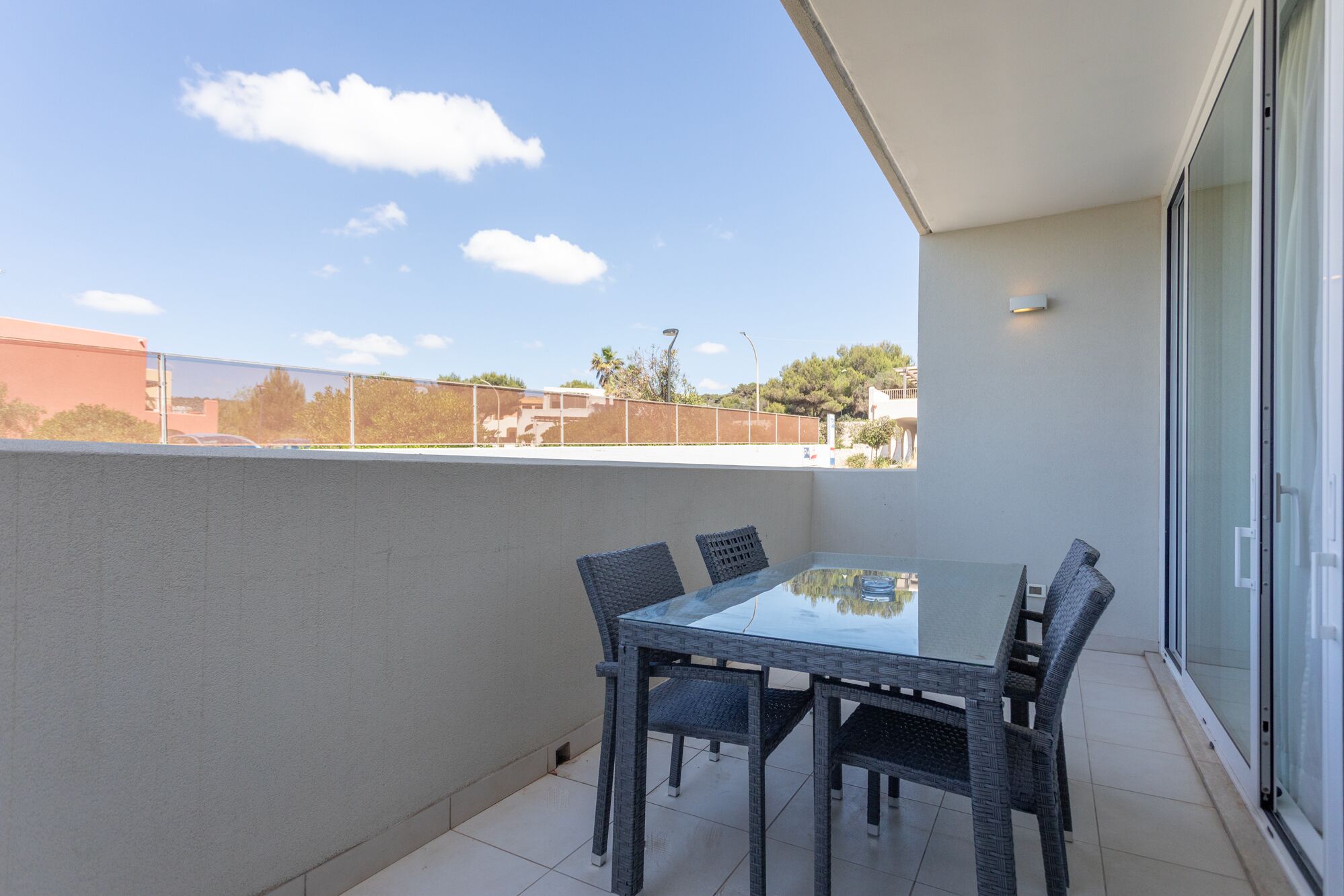 A balcony with a glass table and wicker chairs, overlooking a residential area with houses and trees.