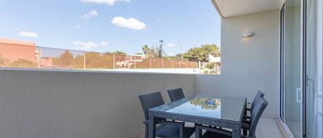 A balcony with a glass table and wicker chairs, overlooking a residential area with houses and trees.