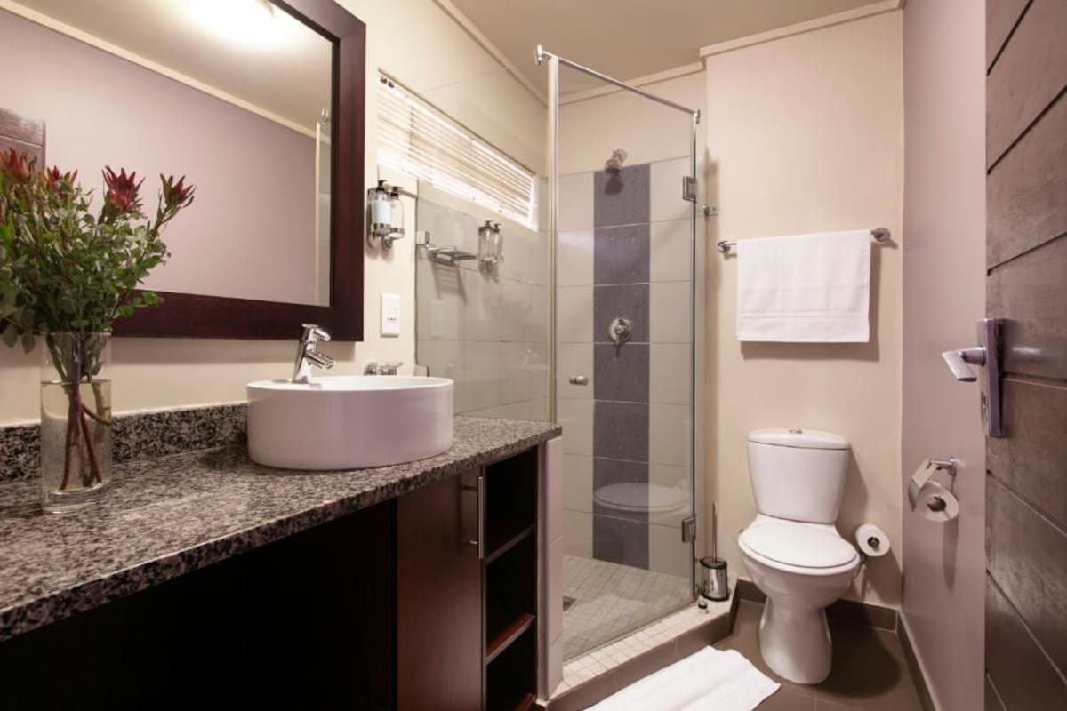 Un baño moderno con encimera de granito, un lavamanos blanco, cabina de ducha de vidrio y un inodoro blanco.