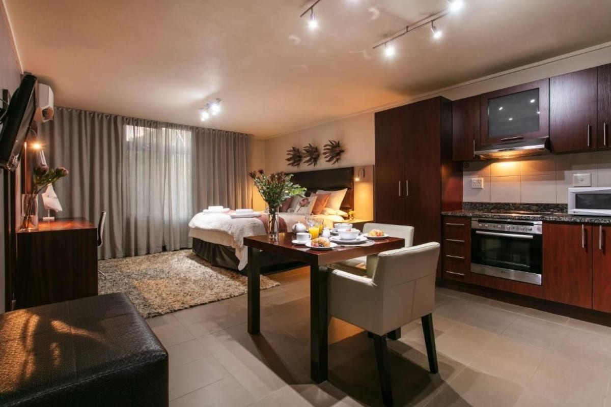 Una habitación de hotel moderna con cama, zona de comedor con mesa y sillas, cocina con armarios de madera oscura y electrodomésticos de acero inoxidable, y un ventanal con cortinas.