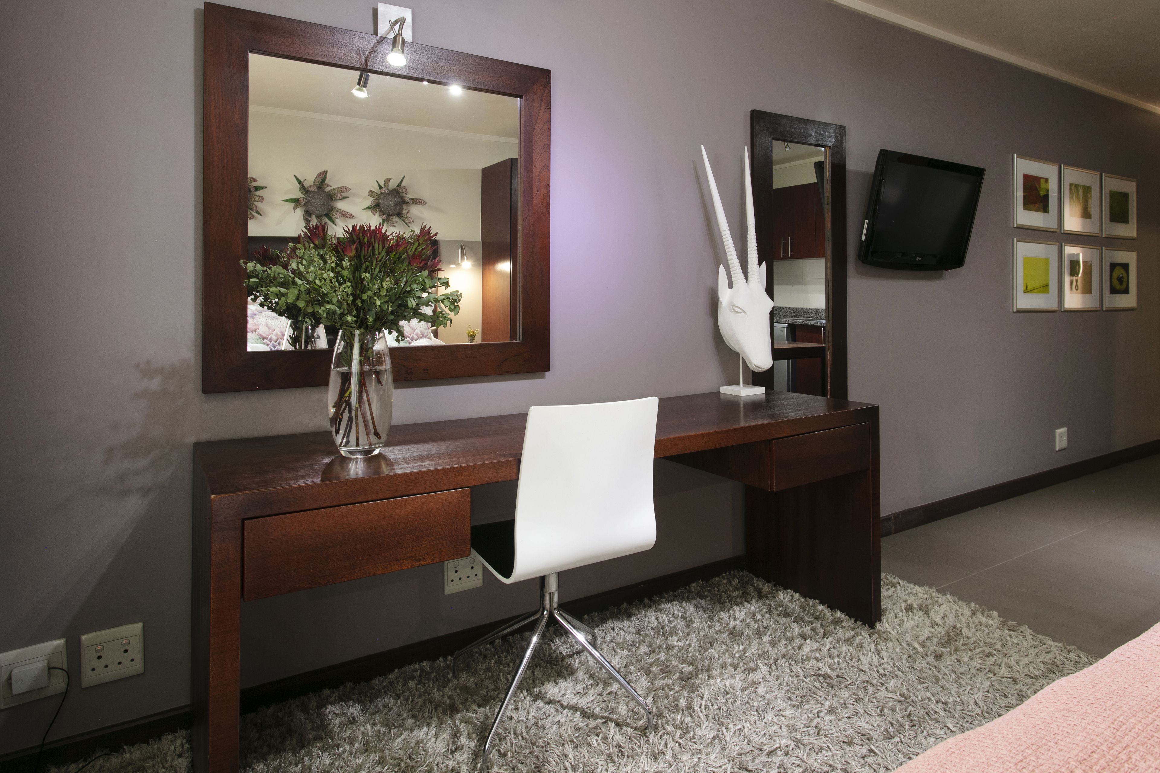 A modern bedroom with a wooden desk, a white chair, a large mirror, a TV, and a decorative skull on the wall.
