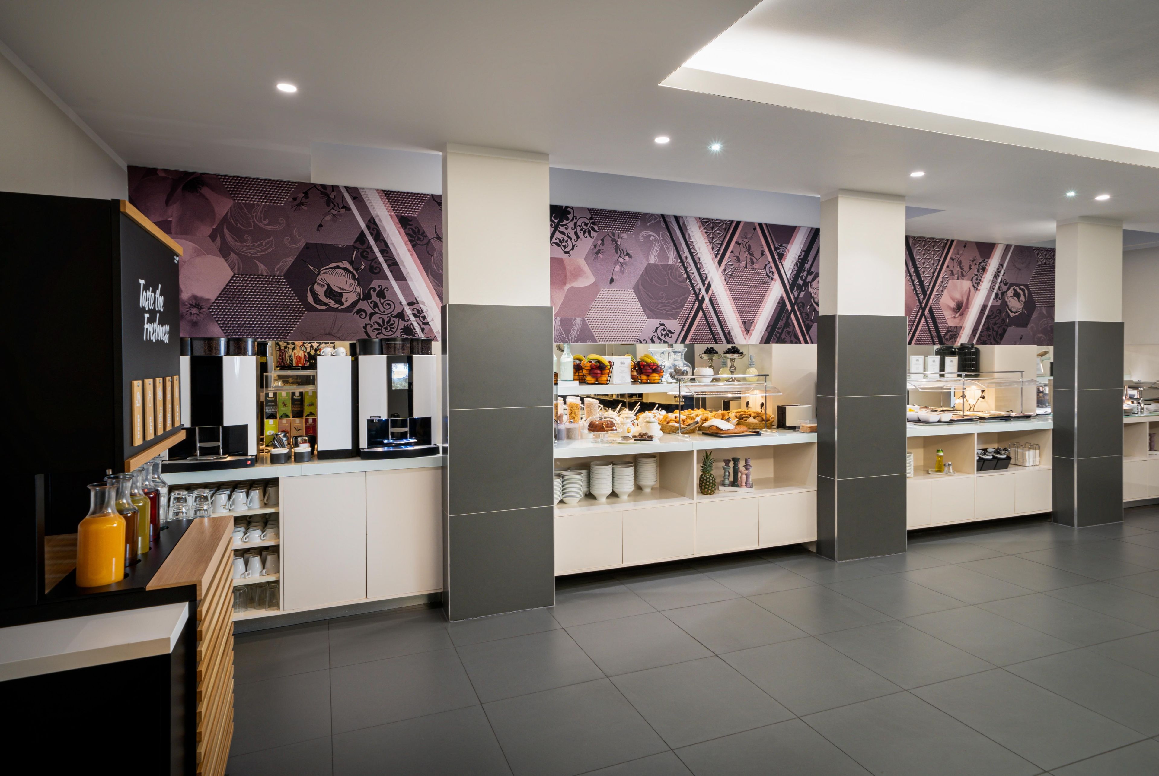 Un hall d’hôtel moderne avec un coin café, un présentoir de fruits et un espace salon.