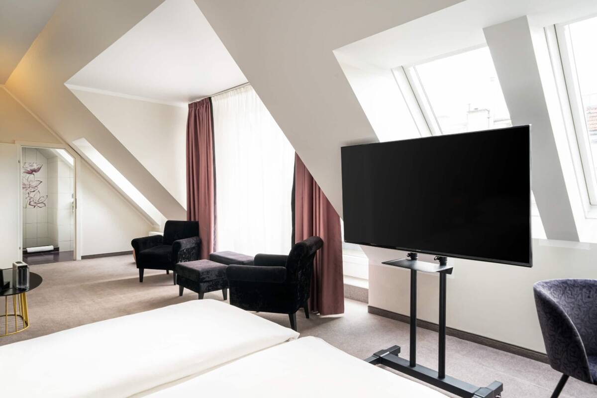 Une chambre d’hôtel avec un grand lit, un téléviseur à écran plat, deux fauteuils, une petite table et une fenêtre avec des rideaux.