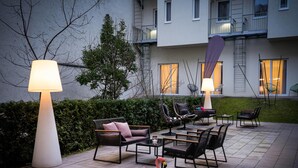 Een terras buiten met modern meubilair, een flamingo standbeeld en een gebouw met balkons op de achtergrond.