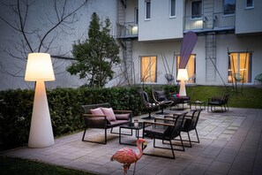 Een terras buiten met modern meubilair, een flamingo standbeeld en een gebouw met balkons op de achtergrond.