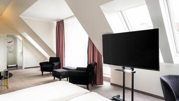 Una habitación de hotel con una cama grande, un televisor de pantalla plana, dos sillones, una mesita y una ventana con cortinas.
