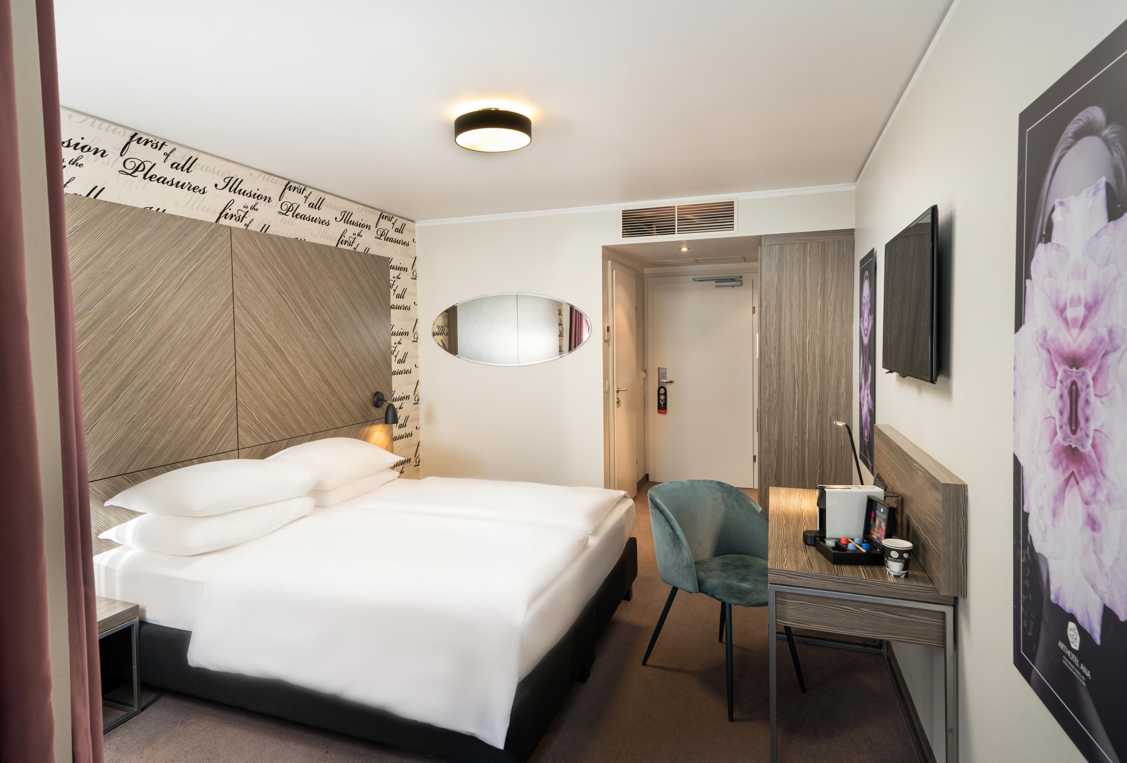 Une chambre d’hôtel comprenant un lit, un bureau avec une chaise, un miroir, une télévision et un mur orné d’une affiche à motifs floraux.