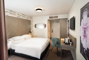 A hotel room with a bed, a desk with a chair, a mirror, a television, and a wall with a floral poster.