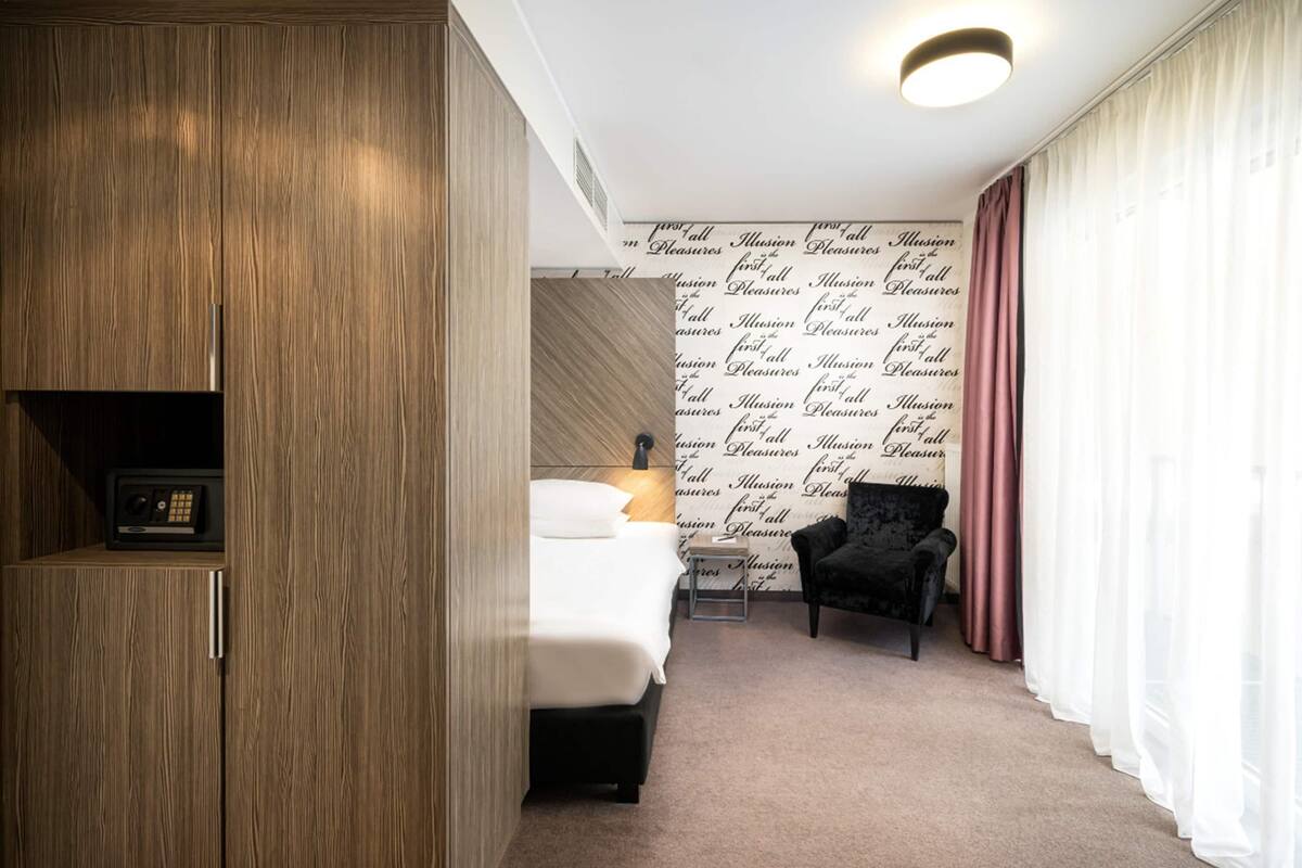 Une chambre d’hôtel avec une armoire en bois, un lit avec une literie blanche, un fauteuil noir, une petite table avec une lampe et un mur orné d’un motif répétitif.