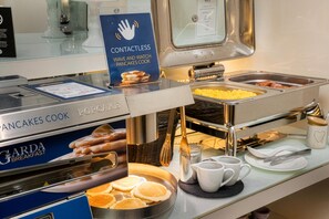 Ein Frühstücksbuffet mit einem Schild für kontaktloses Servieren, einer Grillplatte für Pfannkuchen und verschiedenen Speisen.