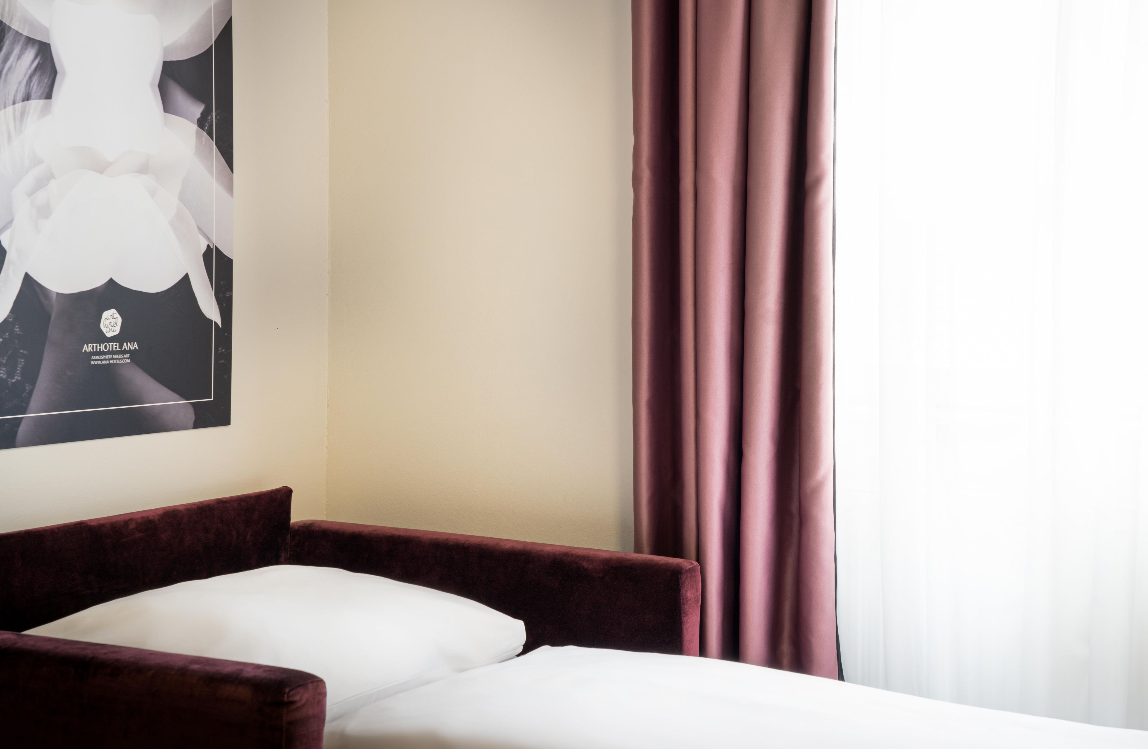 Een hotelkamer met een bed, een rode fluweelstoel, een poster aan de muur en een raam met gordijnen.
