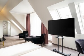 Une chambre d’hôtel avec un grand lit, un téléviseur à écran plat, deux fauteuils, une petite table et une fenêtre avec des rideaux.