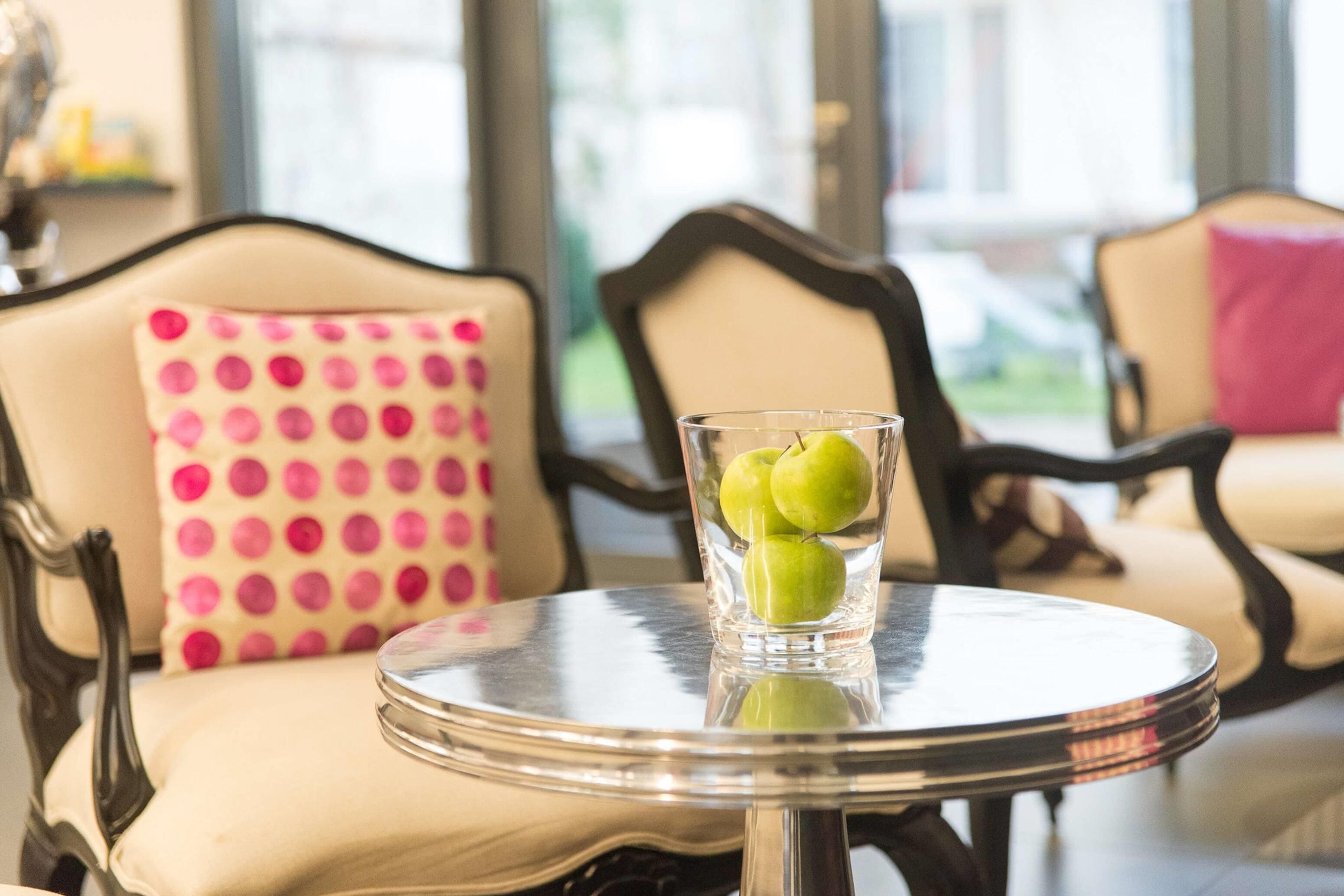 A round table with a glass containing three green apples, a polka-dotted pillow, and a chair with a dark frame.