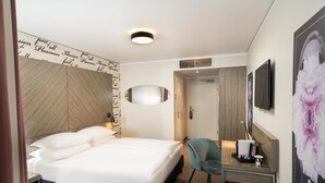 Une chambre d’hôtel comprenant un lit, un bureau avec une chaise, un miroir, une télévision et un mur orné d’une affiche à motifs floraux.