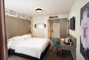 Une chambre d’hôtel comprenant un lit, un bureau avec une chaise, un miroir, une télévision et un mur orné d’une affiche à motifs floraux.
