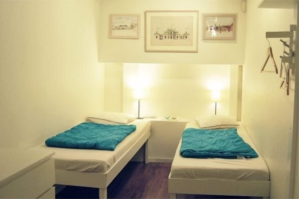 Une petite chambre d’hôtel composée d’une seule pièce, avec deux lits simples, chacun recouvert d’une couverture turquoise. On y trouve deux lampes de chevet, une petite table et une étagère fixée au mur, ornée de tableaux encadrés.