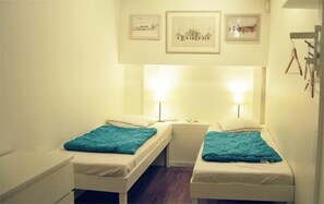 A small, single-room hotel room with two single beds, each with a teal blanket. There are two bedside lamps, a small table, and a wall-mounted shelf with framed pictures. - City Lodge Stockholm AB (Stockholm)