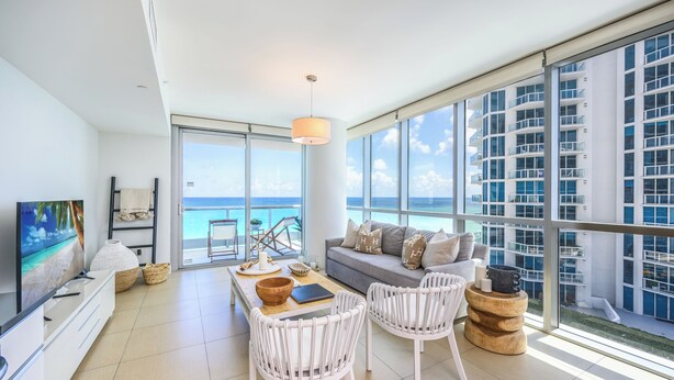 A modern living room with a large window overlooking the ocean, a sofa, white wicker chairs, and a dining table with a bowl and a book. - Monte Carlo by Miami Vacations (Miami Beach)