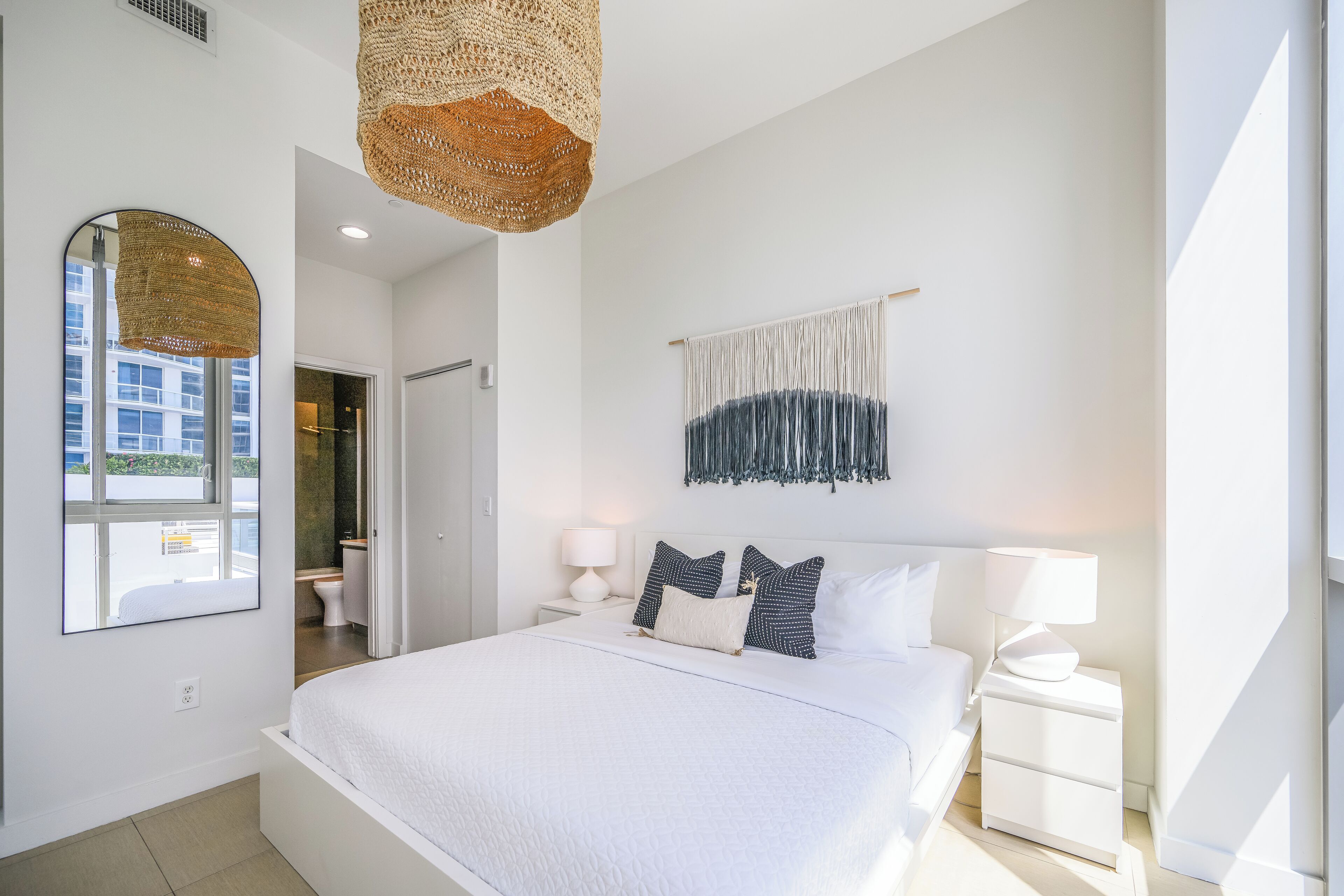 Ein modernes Schlafzimmer mit einem großen Bett, Nachttischen, einer Pendelleuchte, einem Spiegel und einem Wandbehang aus Makramee.