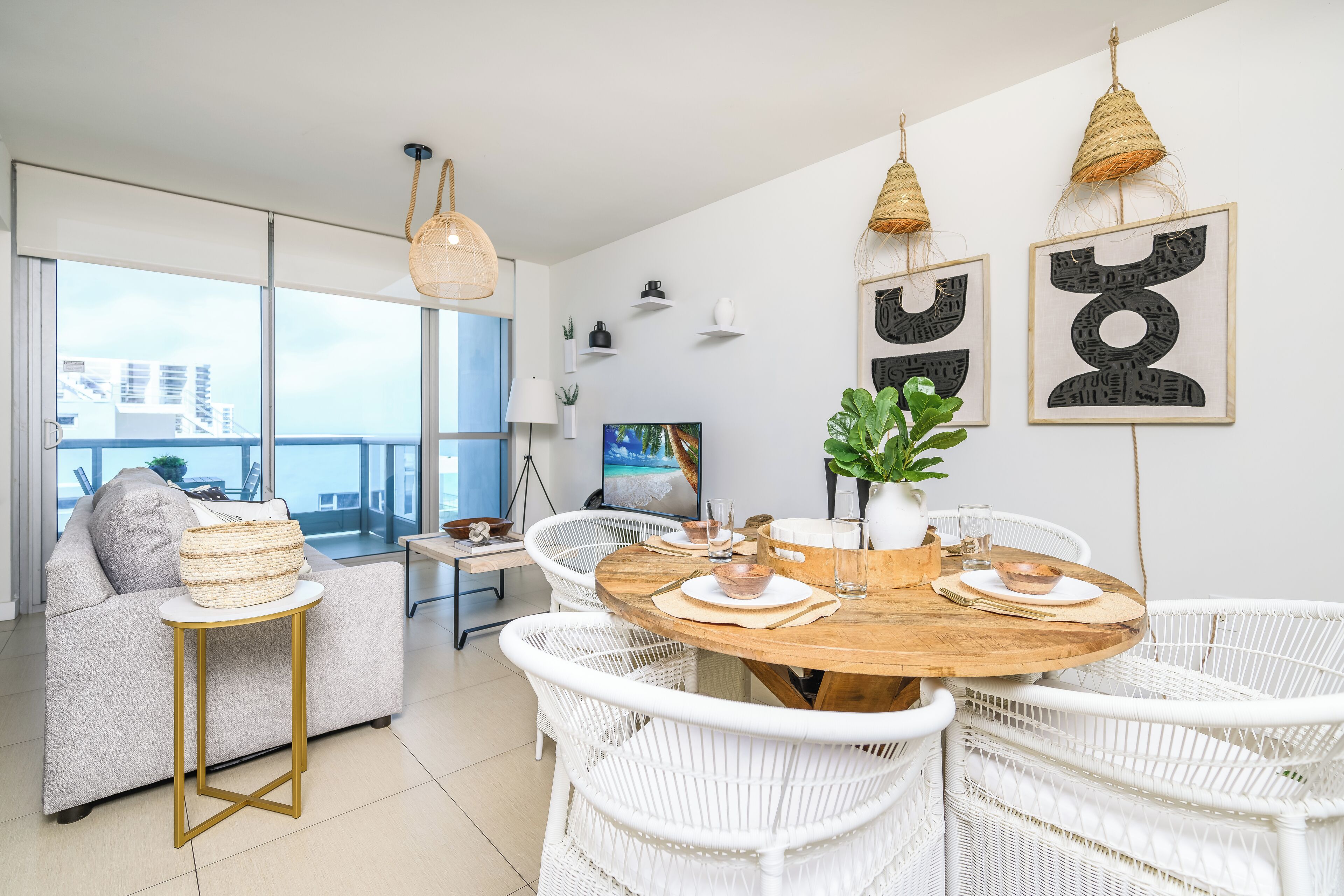 A modern living room with a round wooden dining table, white wicker chairs, a grey sofa, and a balcony with a view of buildings.