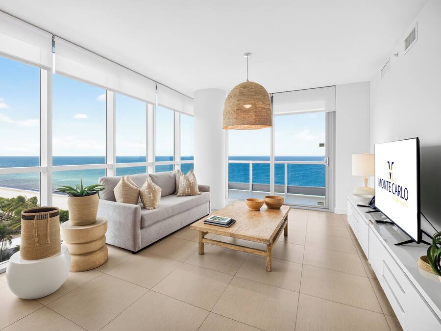 A modern living room with a large window overlooking the sea, a sofa with cushions, a coffee table, and a television.