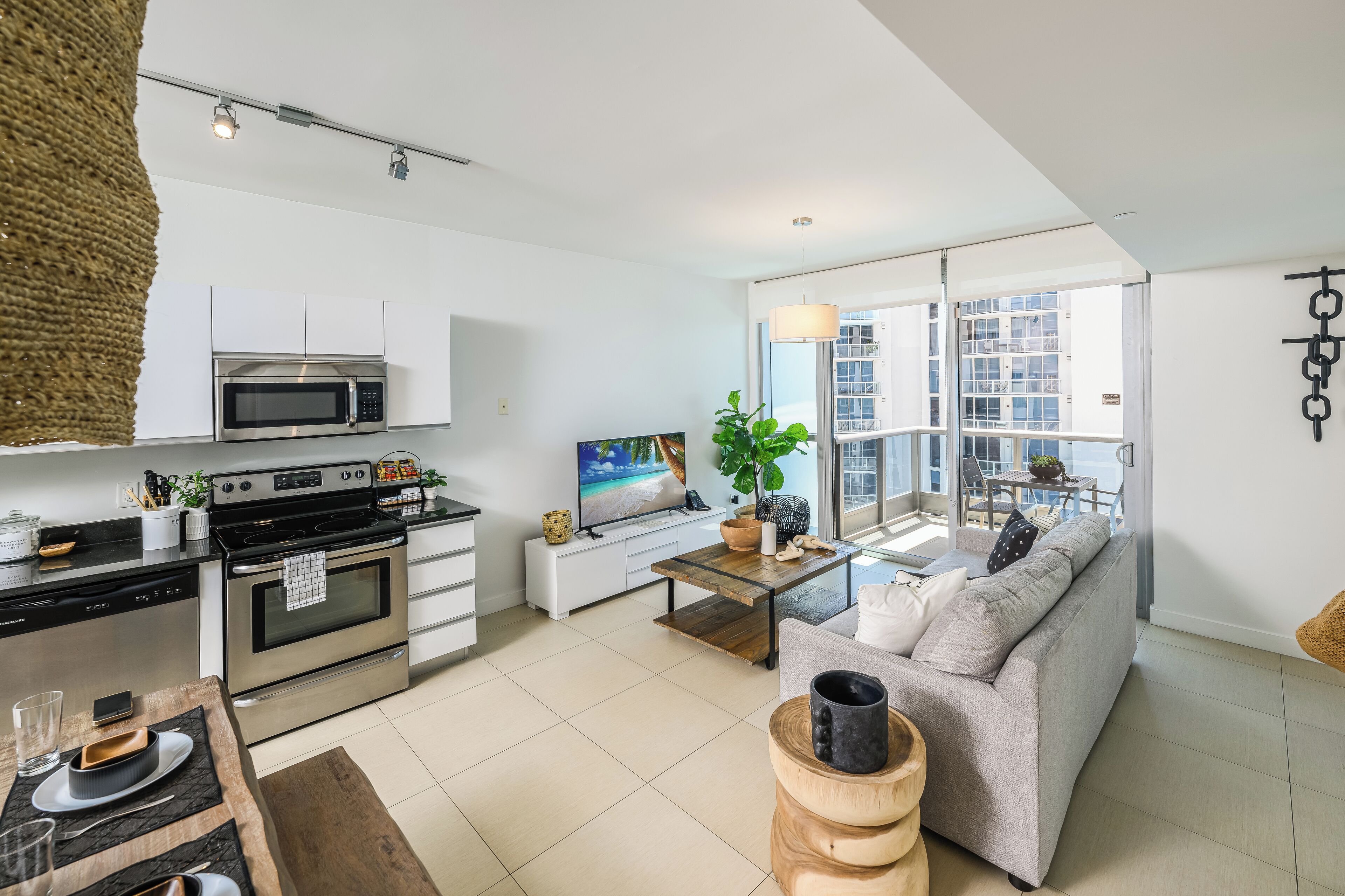 Eine moderne Küche mit Edelstahlgeräten, ein Essbereich mit Holztisch und -stühlen, ein graues Sofa und ein großes Fenster mit Blick auf die Stadt.