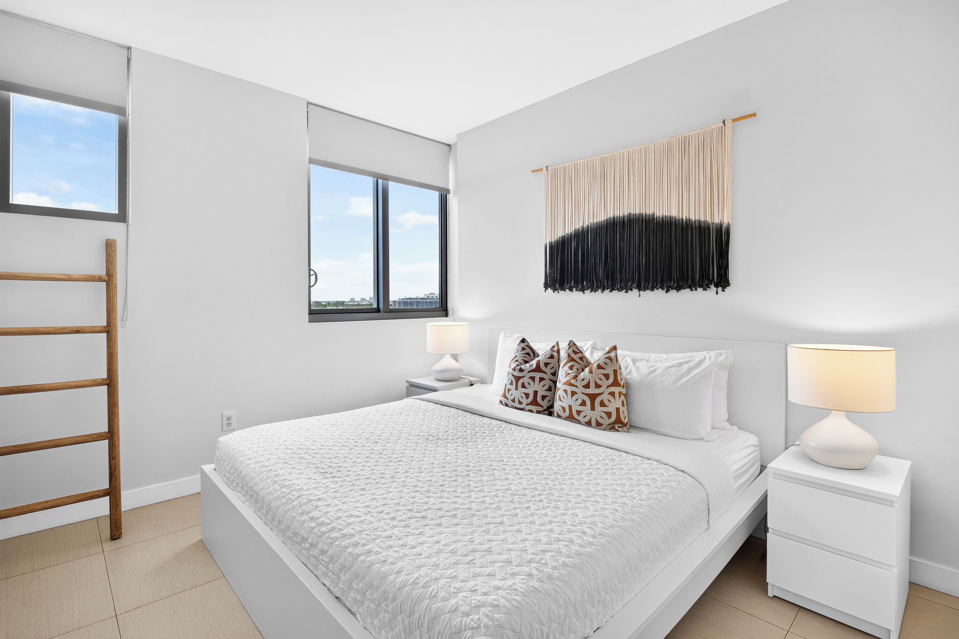 A modern bedroom with a large bed, two bedside tables, a hanging macrame wall decoration, and a window with a city view.
