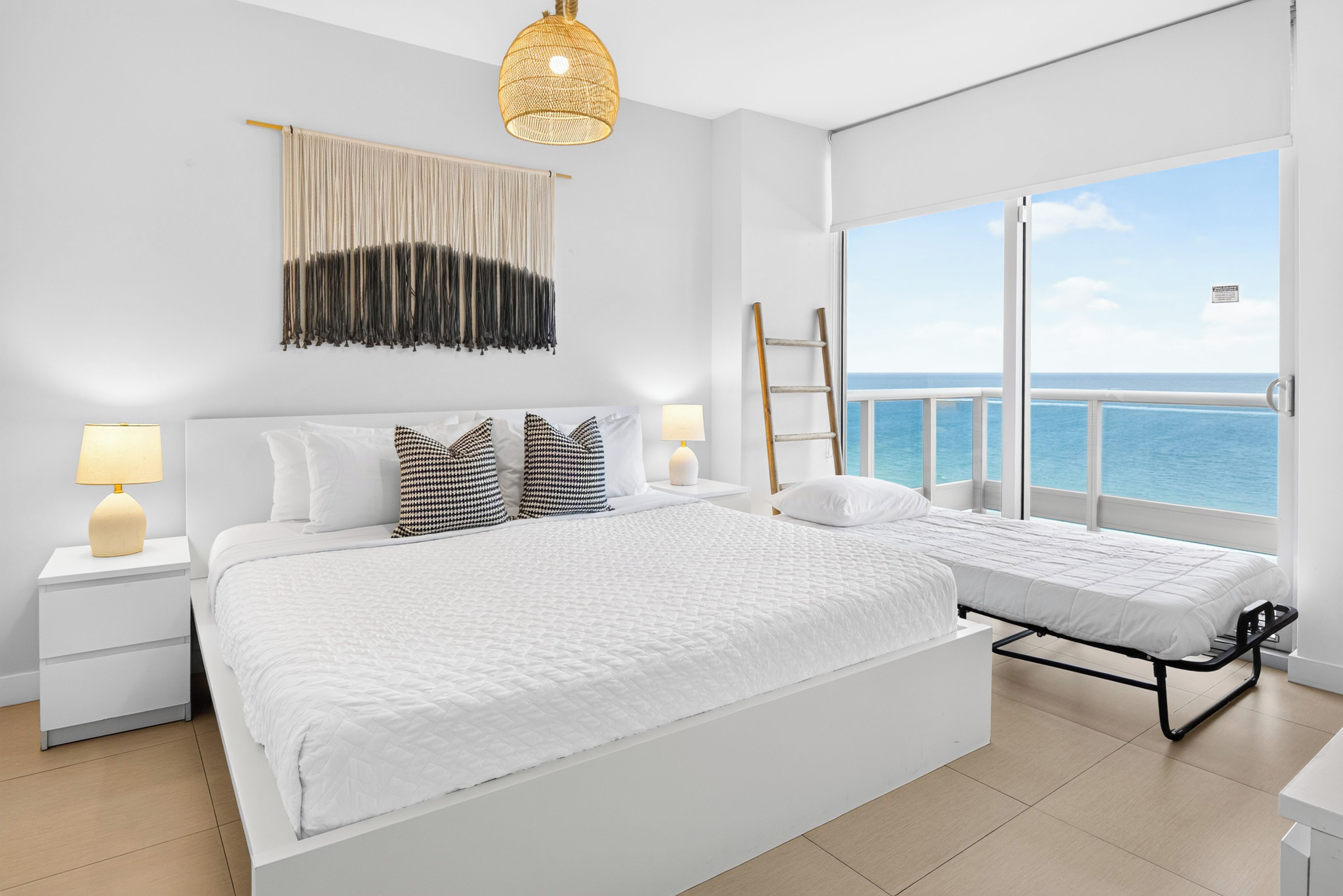 Un dormitorio con una cama grande, dos mesitas de noche con lámparas, un candelabro, vista al mar y un balcón con barandilla.