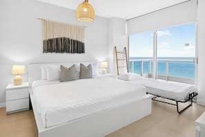 Een slaapkamer met een groot bed, twee nachtkastjes met lampen, een hanglamp, uitzicht op zee en een balkon met een hekwerk.