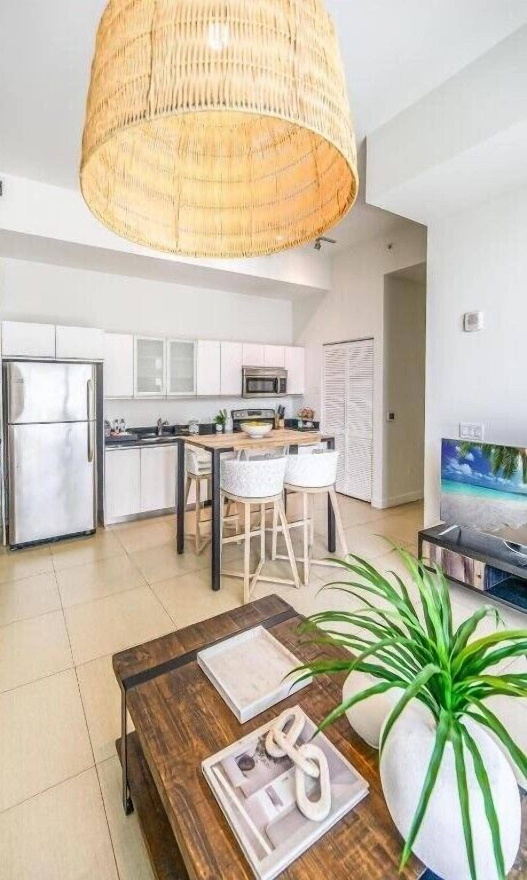 A modern kitchen with white cabinets, a stainless steel refrigerator, and a microwave. There is a dining area with a wooden table and two white chairs. A large wicker lamp hangs from the ceiling, and a potted plant is on the floor.