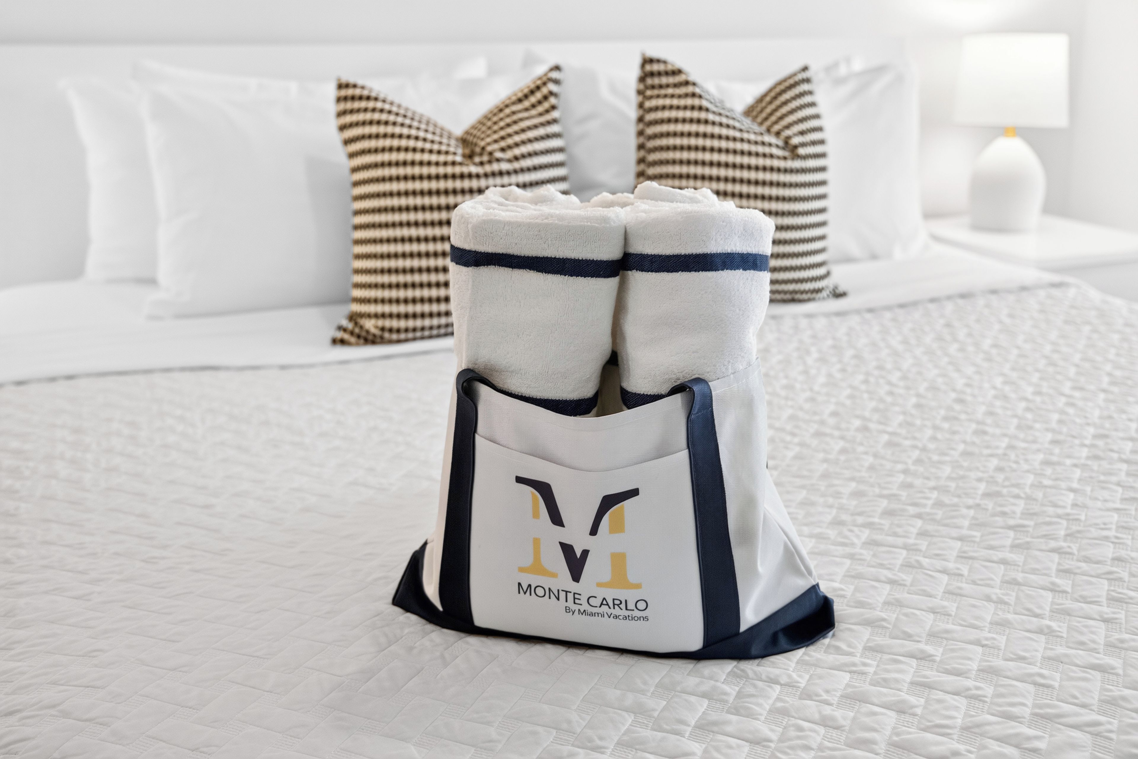 Una cama bien hecha con una colcha blanca acolchada, un almohadón estampado en cuadros y un par de pantuflas blancas en una bolsa etiquetada 'Monte Carlo by Miami Vacations'.