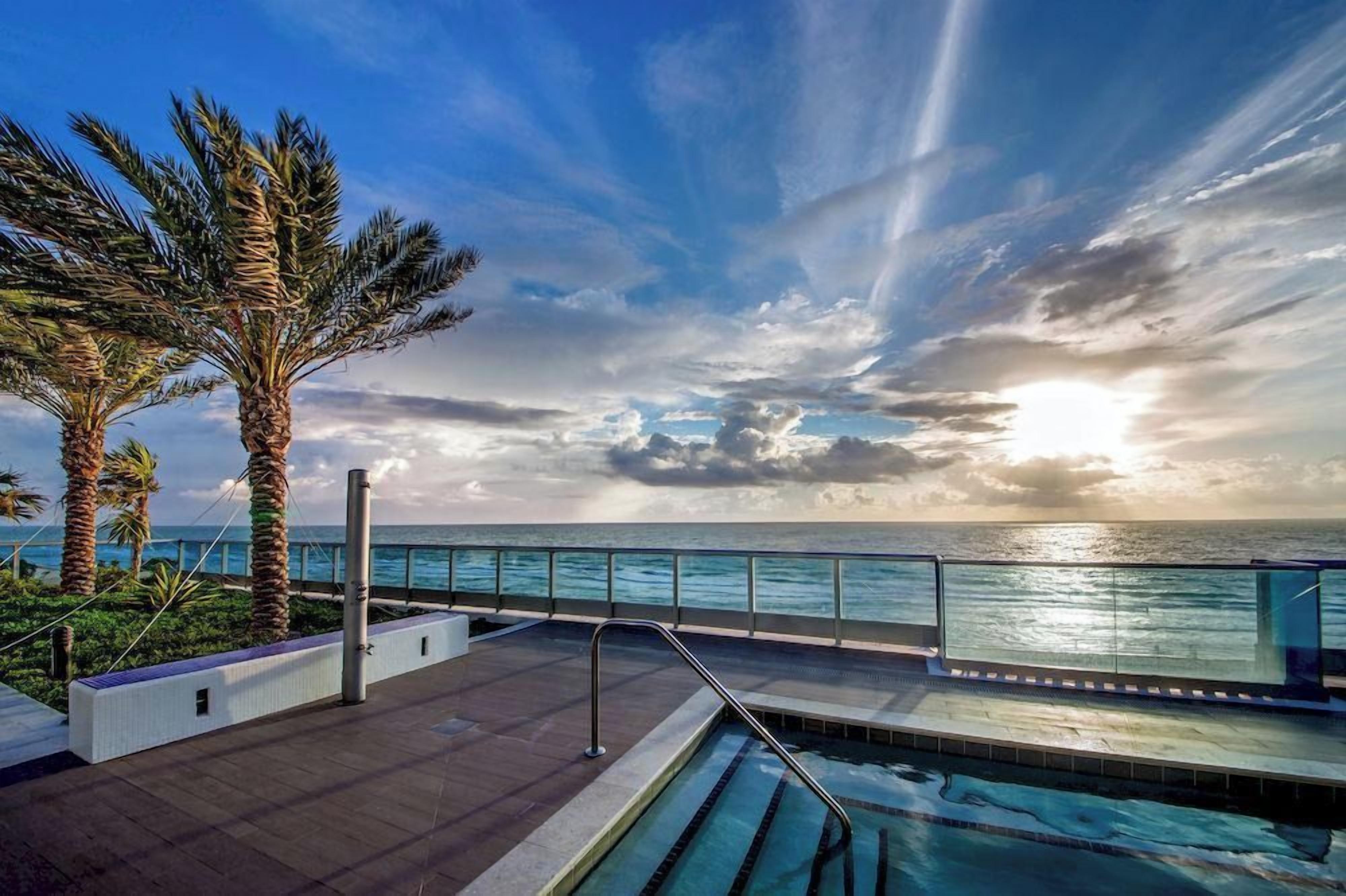 Un área de piscina con vistas al océano, palmeras y un cielo despejado.