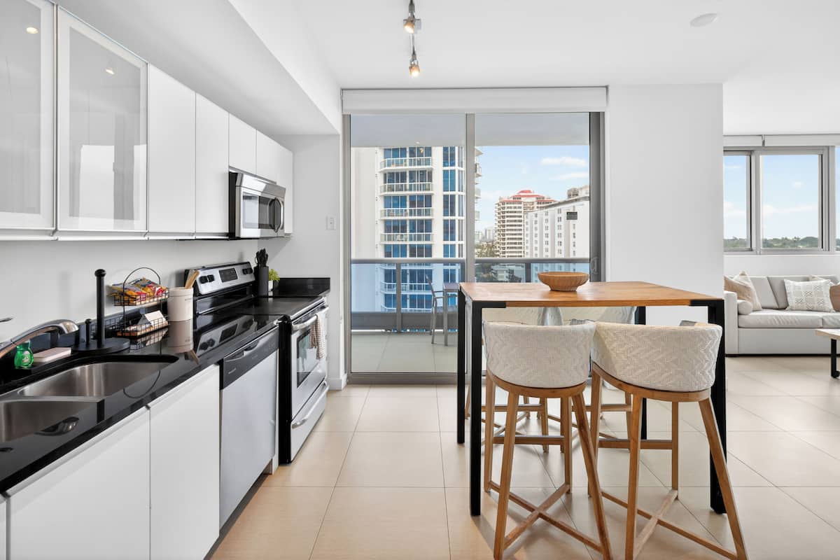 A modern kitchen with a dining area, a large window with a city view, and a balcony.