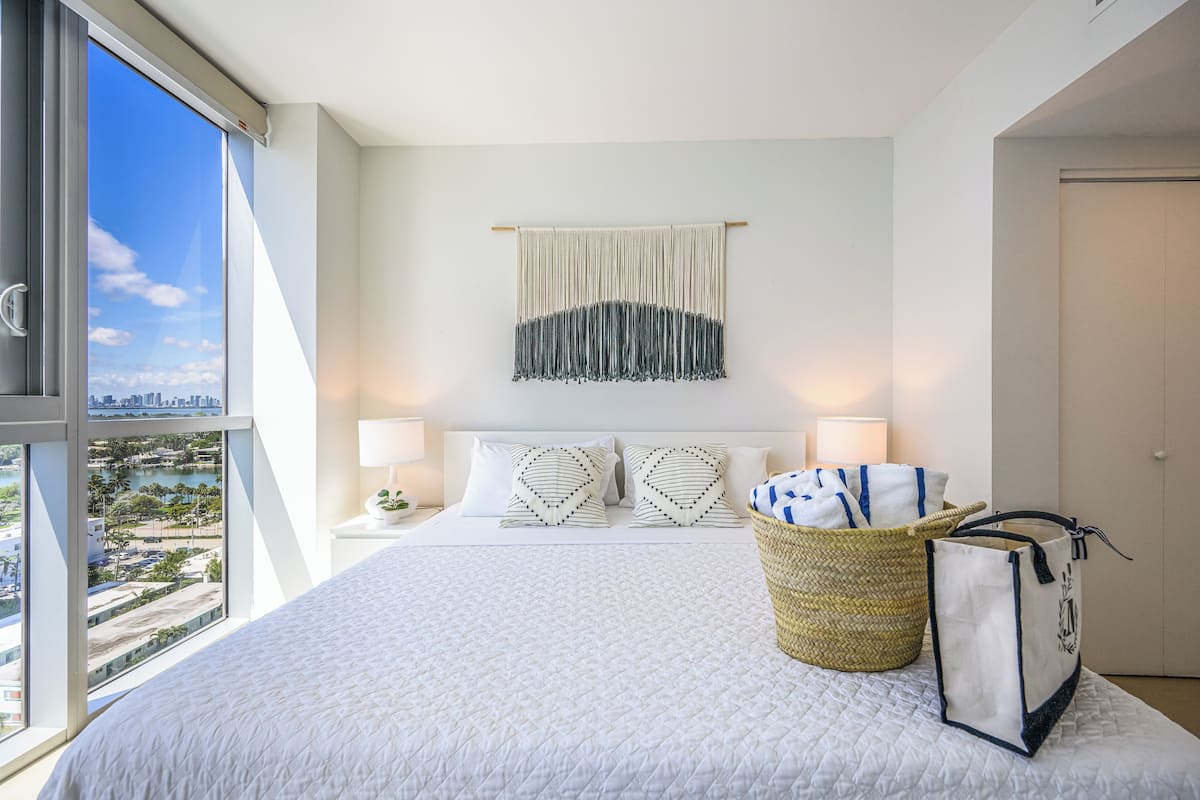 A hotel room with a large bed, a view of the city, a macrame wall hanging, and a wicker basket with towels.