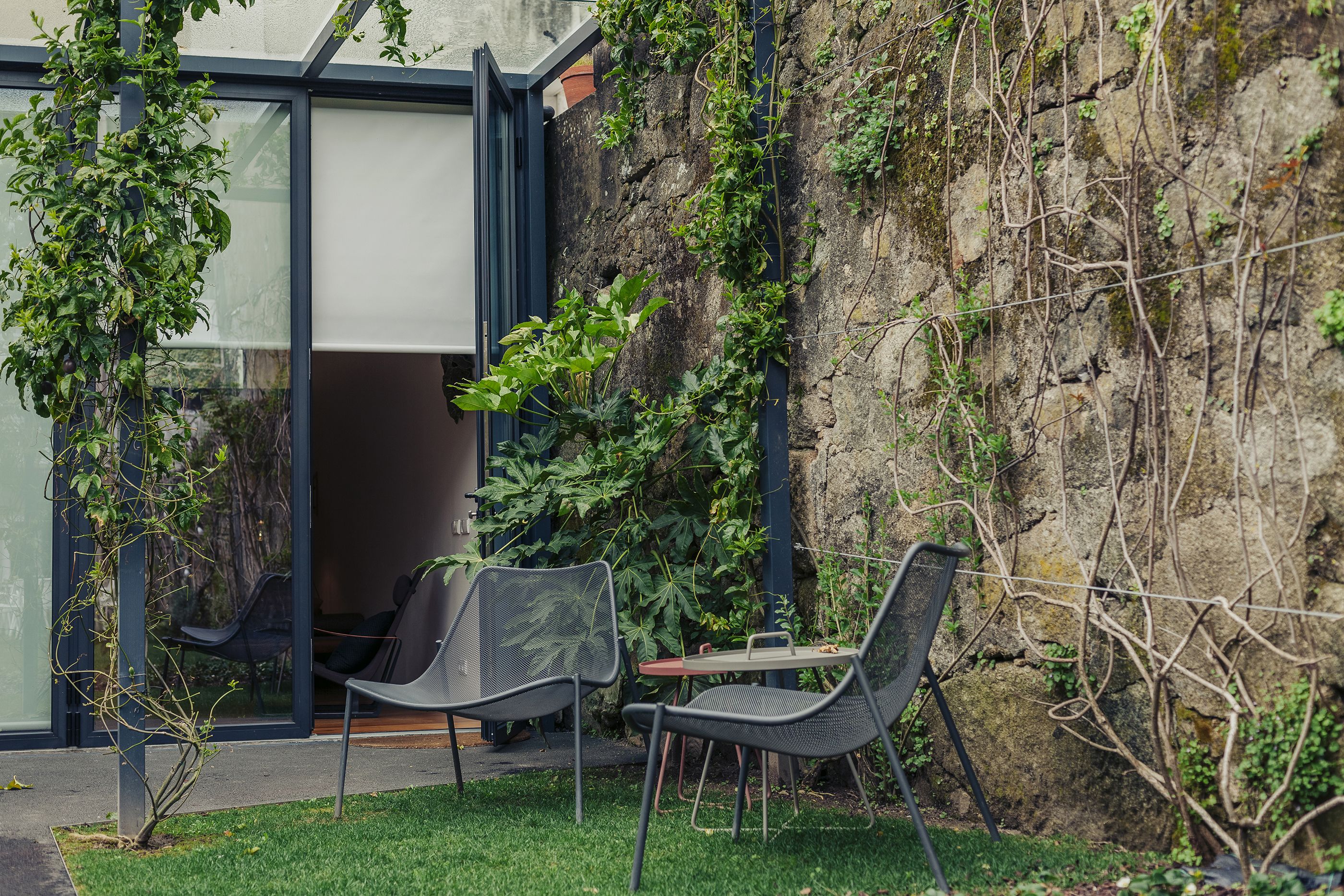 An outdoor seating area with two chairs and a table, surrounded by greenery and a stone wall.