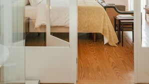A modern hotel room with a glass partition, a wooden floor, a bed with white bedding, a small table, and a hanging light fixture. - Rosa Et Al Townhouse (Porto)
