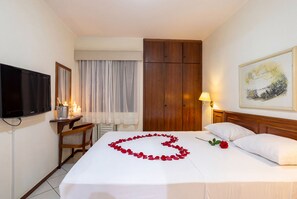 Quarto de hotel com uma cama, televisão, escrivaninha com luminária, guarda-roupa de madeira e um quadro na parede.