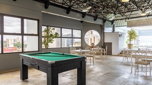 A pool table in a room with large windows, a dining area, and a view of buildings. - Park Tower Campinas Residence by Nacional Inn (Campinas)