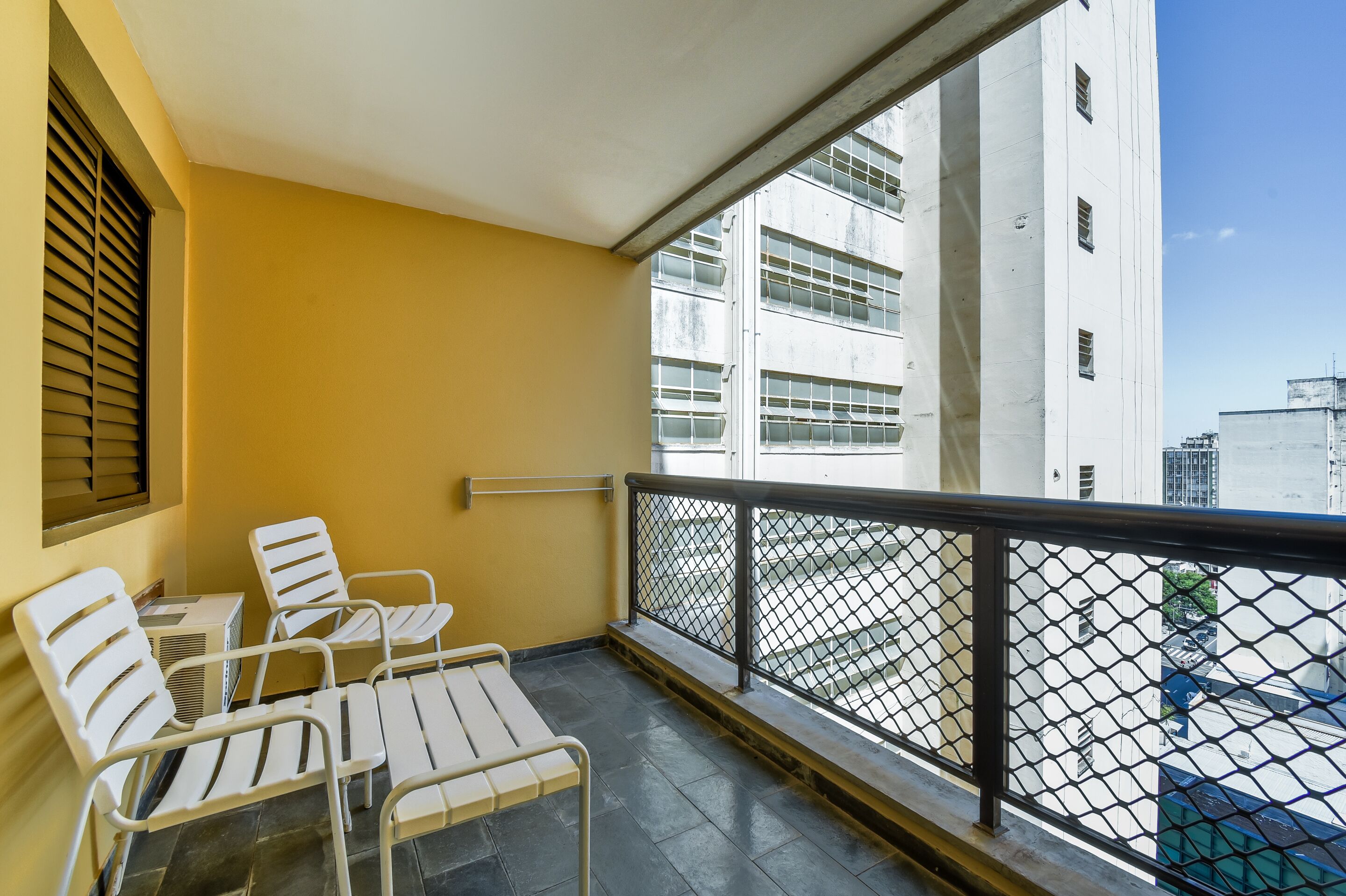 A balcony with white chairs and a table, a metal railing, and a view of a white building with multiple windows.