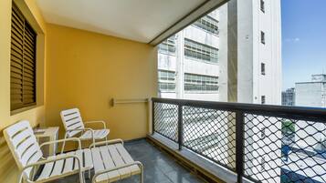 Varanda com cadeiras e mesa brancas, guarda-corpo de metal e vista para um edifício branco com várias janelas.