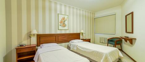 Habitación de hotel con dos camas, cabecero de madera, mesita de noche con lámpara, escritorio con silla, ventana con persiana y espejo fijado a la pared.