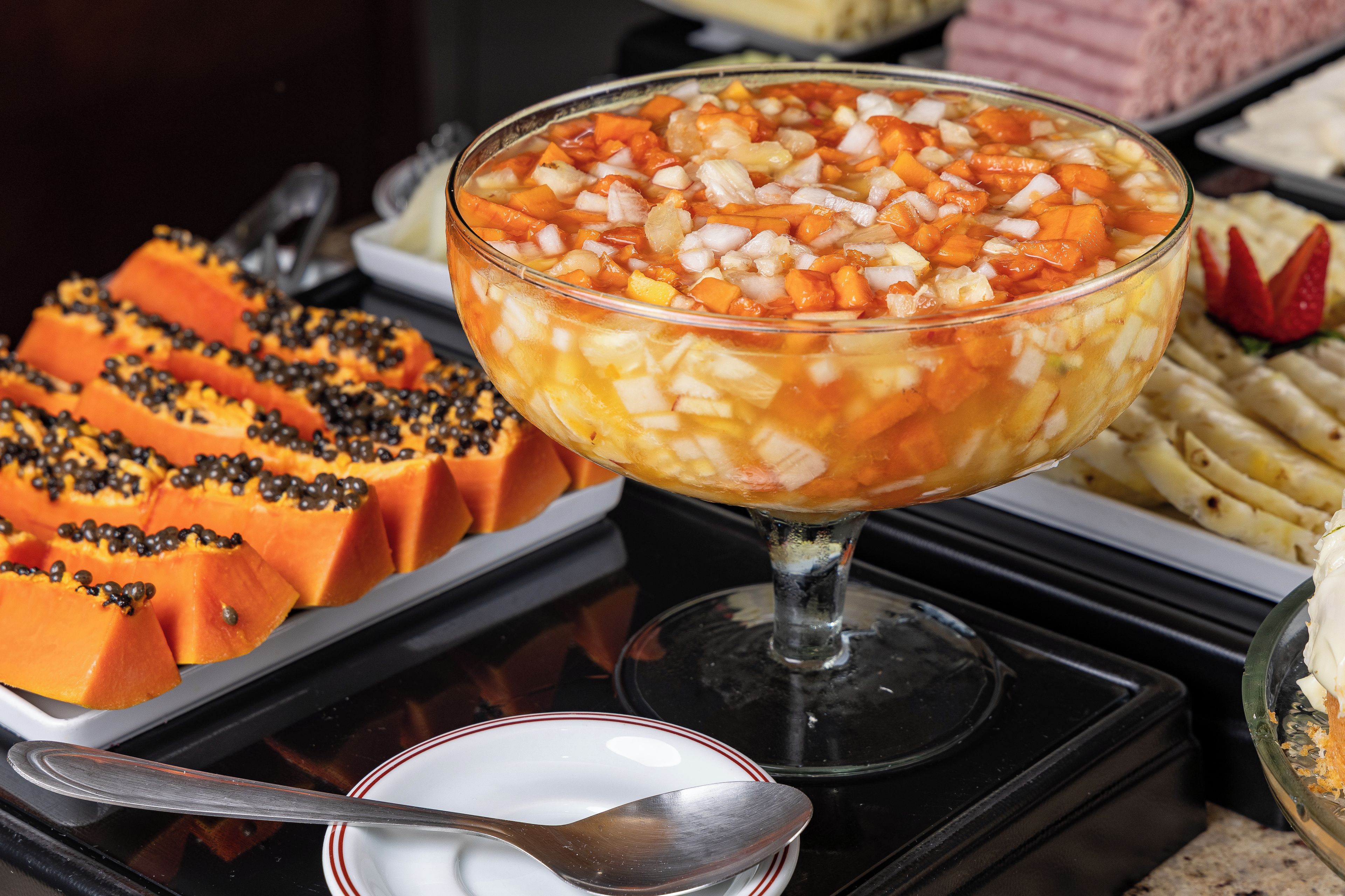 Buffet variado com diversos pratos, incluindo uma grande saladeira de salada de frutas, mamão fatiado e outros alimentos preparados.